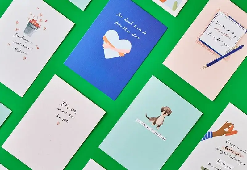 A collection of colorful greeting cards arranged on a bright green background. The cards feature various designs and handwritten-style messages, including illustrations like a bucket of hearts, a dog, and a hand gesture